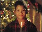 christmas Devotion < young boy in plaid pajamas holding a single lit candle with christmas tree in background